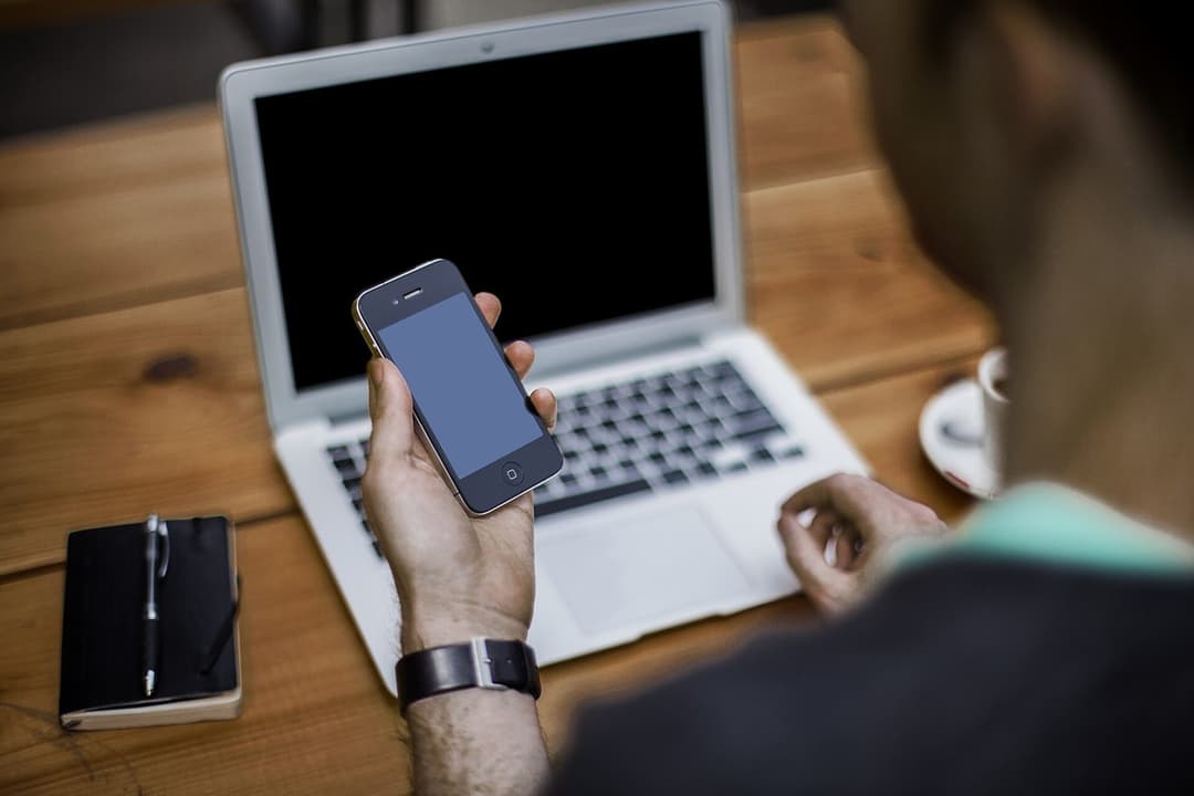 A man holding a smartphone at a laptop